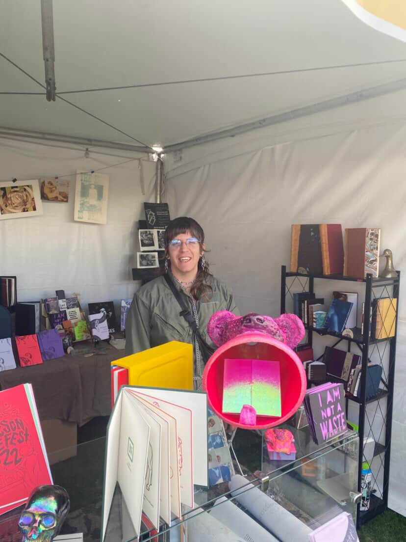 Emiland Kray standing behind a table display inside a white tent, smiling, surrounded by their colorful handmade books and art pieces arranged on shelves and a table.