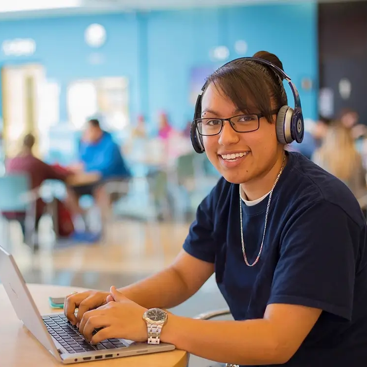 A student smiles at the camera sitting in a cafeteria on a laptop