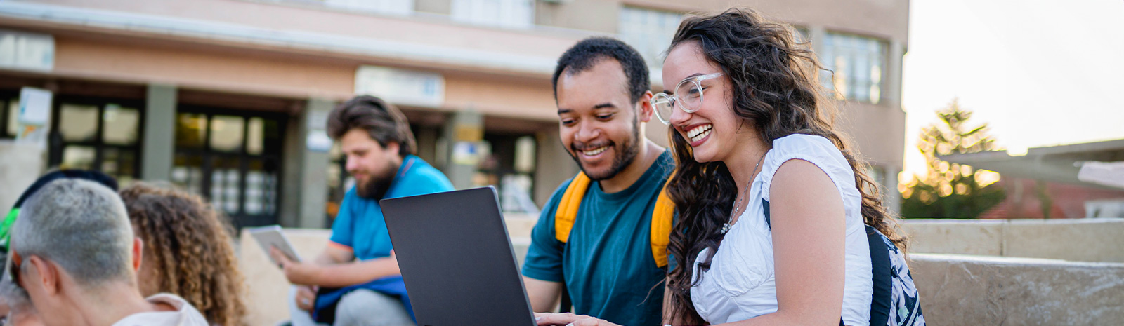 Two students sit in a courtyard laughing while looking at a laptop