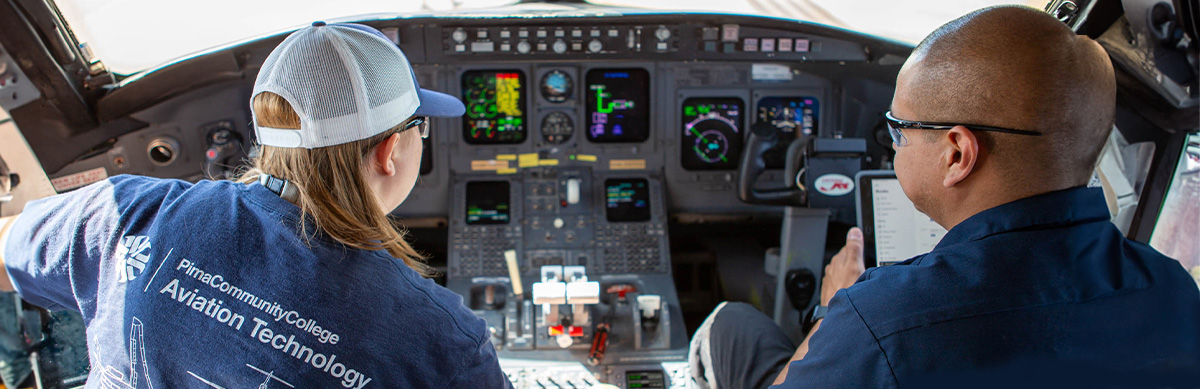 Two aviation students sit in the cockpit of a commercial airplane working on controls