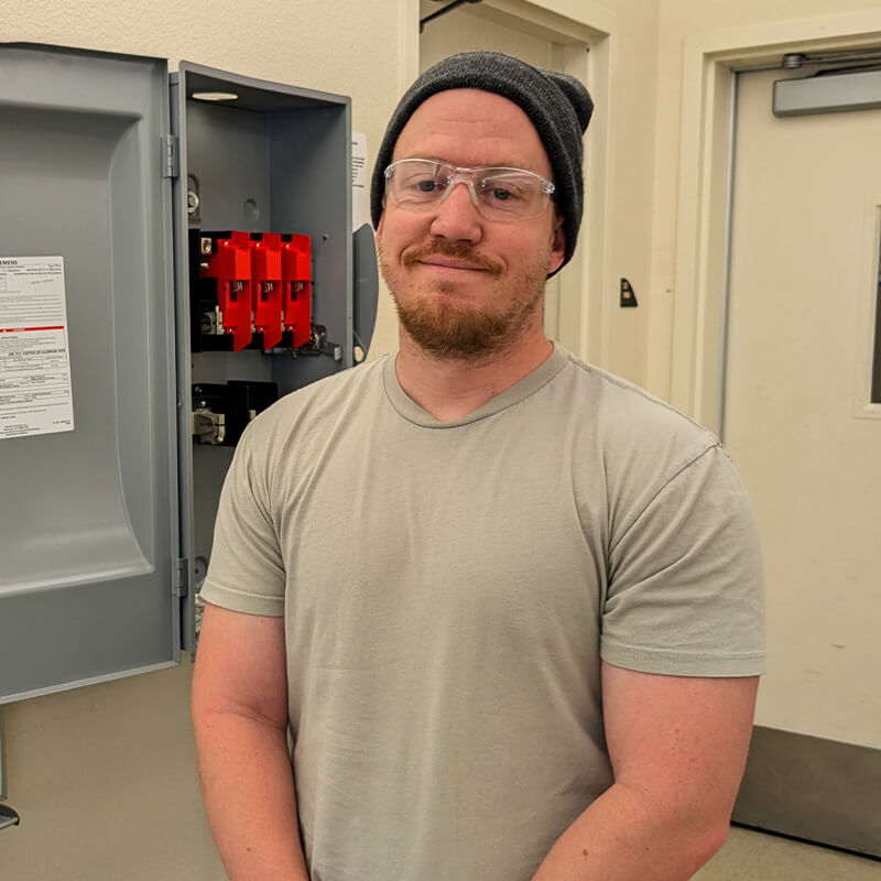 Thomas stands smiling in front of an open electrical box